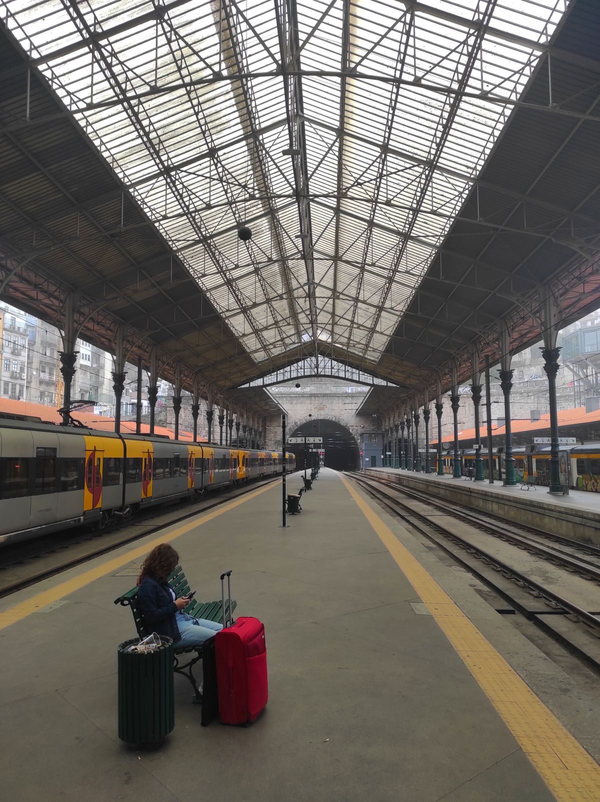 Voyageuse assise avec une valise rouge sur un quai de gare sous une grande verrière, un train à quai sur la gauche